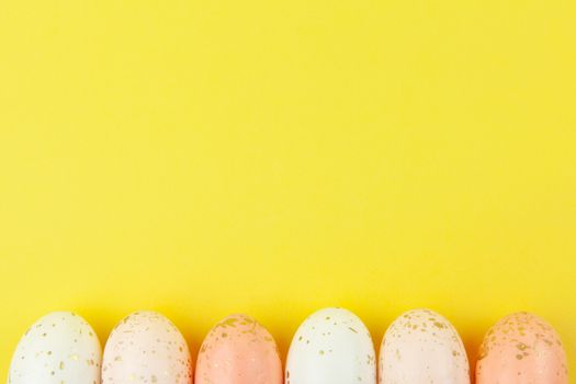 Creatively painted eggs in pastel colors decorated with gold leaf are arranged in row along bottom edge on yellow background, copy space. Happy Easter DIY concept. Flat lay. Horizontal.