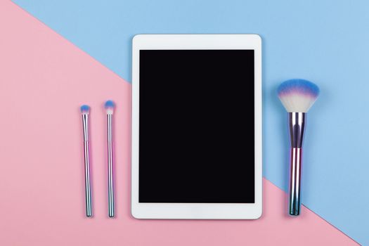 Modern working space of visagiste, top view. Tablet, makeup brushes on two-color background pink and blue, copy space, flat lay. Small business, work from home, using technology concept. Horizontal.