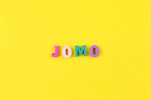 Abbreviation word JOMO in multicolored wooden letters on pastel yellow background. JOMO - Joy Of Missing Out. Opposition, choice, social problem, digital detox. Flat lay, copy space, minimalism style.