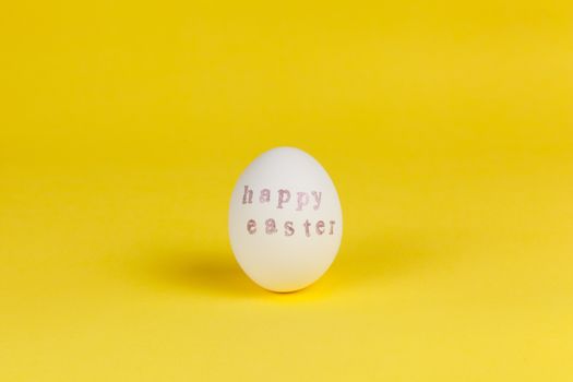 One white chicken egg with words Happy Easter written with help of stamps and pink metallic paint on yellow background, copy space. DIY, festive concept. Side view. Horizontal.