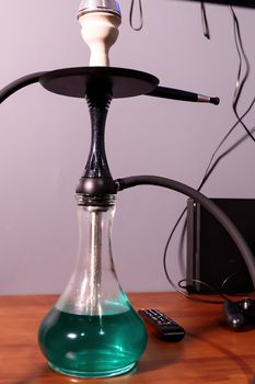 Black matte hookah with clear water with a black hose on the table next to the remote control in the lounge bar on a light background. Hookah cafe leisure concept.