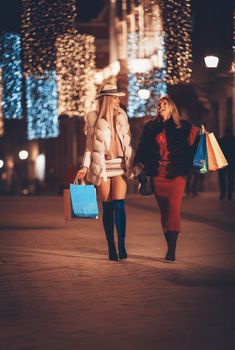 Young beautiful cheerful female friends with colorful shopping bags having fun and walking in the city street at night at Christmas time.