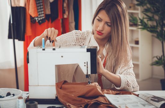 Young beautiful pensive girl sew with a sewing machine. In the background you can see hanging garments.