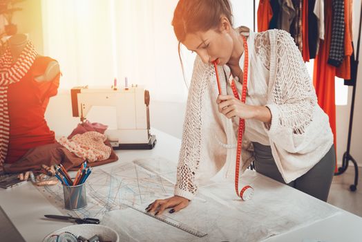 Young pensive female fashion designer with tape measure draped over the neck drawing on sewing pattern with a ruler.