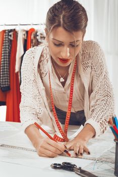 Young female fashion designer with tape measure draped over the neck drawing on sewing pattern with a ruler.