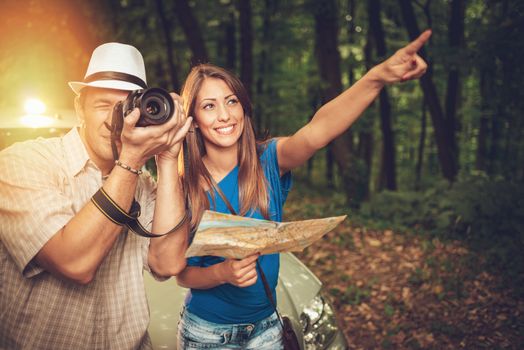 Young travelers standing before a car in the forest. Young man taking a photo with digital camera. Girl holding map and pointing destination. 