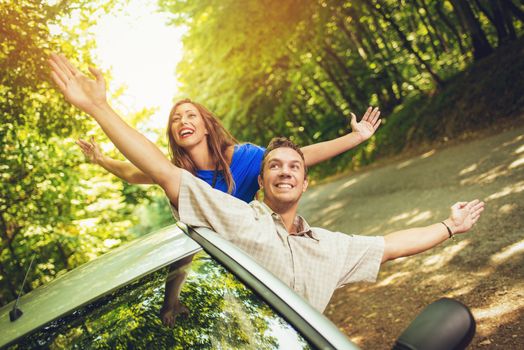 Cheerful travelers on summer travel vacation leaning out of a car window with arms raised.