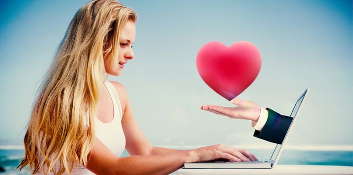 Composite image of pretty blonde using her laptop at the beach with hand holding heart