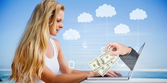 Composite image of pretty blonde using her laptop at the beach with hand holding money