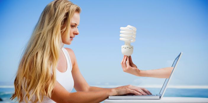 Composite image of pretty blonde using her laptop at the beach with hand holding bulb