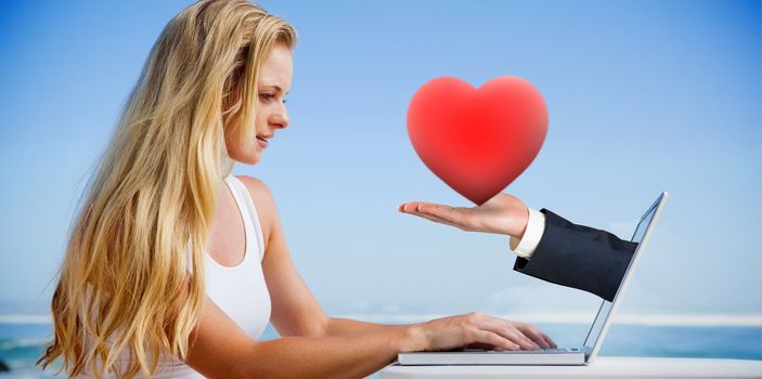 Composite image of pretty blonde using her laptop at the beach with hand holding heart
