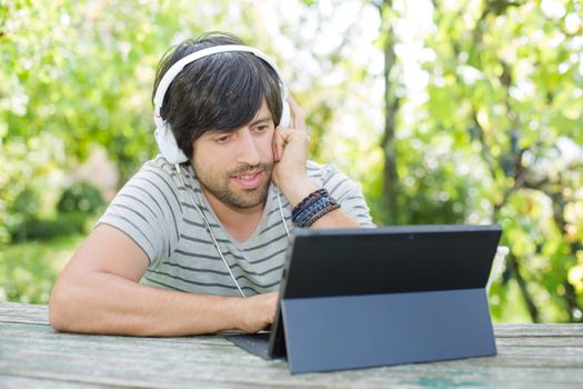 young man working with a tablet pc listening music with headphones on a wooden table, outdoor