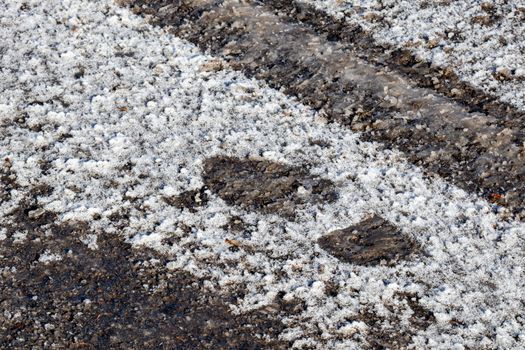 The footprint left behind by a boot has been impressed in a thin layer icy snow on the ground in a parking lot. The paved asphalt is seen within it, and in a parallel tire track.