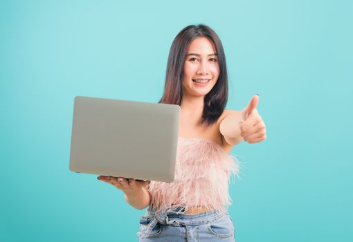 Smiling face portrait asian beautiful woman holding laptop computer and giving thumb up on blue background, with copy space for text