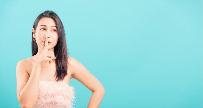 Smiling face Asian beautiful woman her asking quiet with finger on lips pointing with hand on blue background, with copy space for text