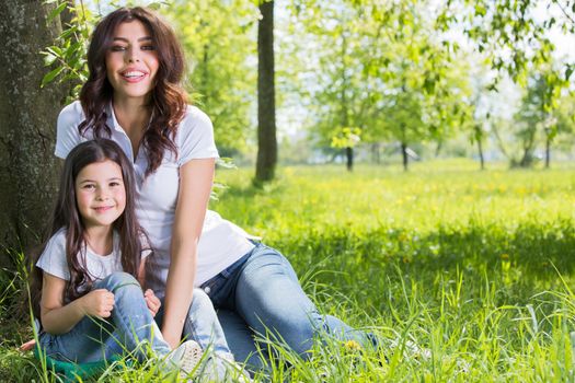 Happy mother with her daughter sitting on grass under the tree in park