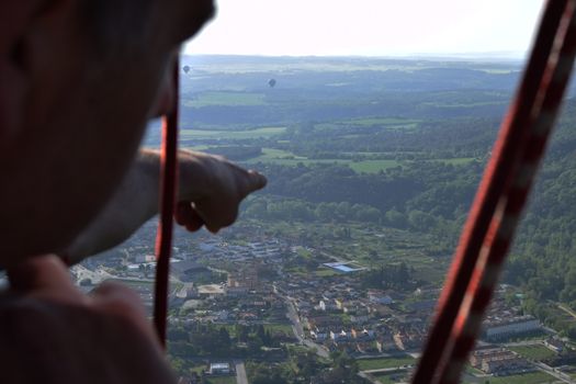 A man pointing from a hot air balloon to the horizon of a beautiful green landscape