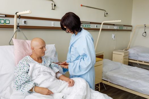 Woman without hair after chemotherapy patient lying at hospital bed feeling sad and depressed worried. A friend came to please her, showing photos on her mobile phone.