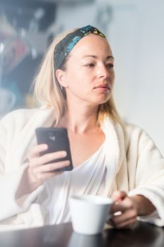 Beautiful caucasian woman at home, feeling comfortable wearing white bathrobe, taking some time to herself, drinking morning coffee and reading news on mobile phone device in the morning.