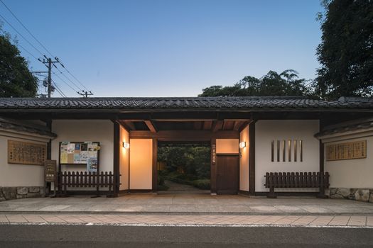 Front gate in longhouse-style of Mejiro Garden with white lacquered walls. This style named nagaya-mon was common in old Edo. Roofed walls made from mud and clay stand on either side, and the walkway is lined with greenery, adding grace and character to the surrounding environment.