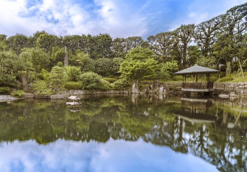 Hexagonal Gazebo Ukimido in the central pond of Mejiro Garden where ducks are resting and which is surrounded by large rocks and stone pagoda under the foliage of  the Japanese pines trees and a variety of momiji maple trees like iroha-momiji, yama-momiji, or oo-momiji. The gazebo has a wickerwork ceiling that beatifully reflects the light from the surface of the water.