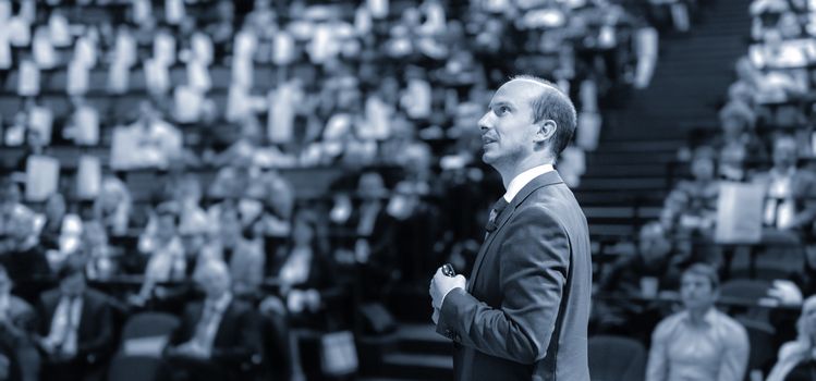 Speaker giving a talk on corporate business conference. Unrecognizable people in audience at conference hall. Business and Entrepreneurship event. Blue toned image.