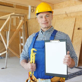 Portrait of worker in blue uniform and yellow hardhat holding adjustable wrench and blank contract at construction site