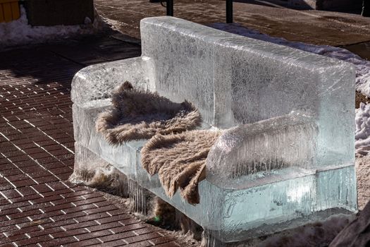 In the winter, an ice bench shaped like a couch or sofa sits on a brick sidewalk. The bench is sculpted out of solid ice and carries warm blankets for pedestrians to rest.