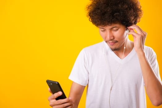 Happy asian handsome man with curly hair he smiling enjoying listening to music on earphones using a mobile smartphone isolate on yellow background