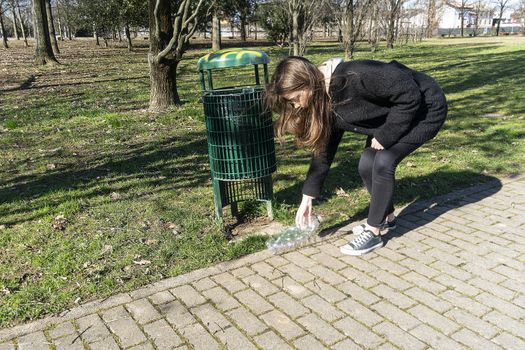 a girl picks up on the ground a plastic bottle and throws it in a waste basket