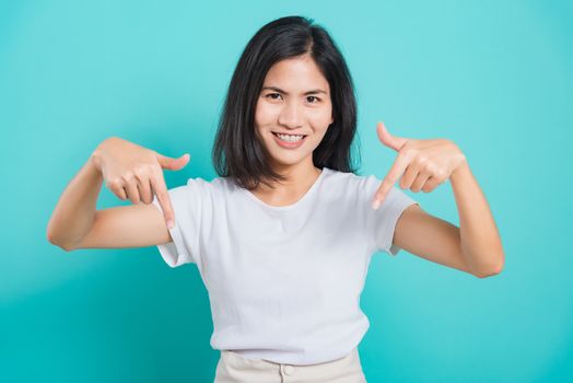 Portrait Asian beautiful young woman standing wear t-shirt, She points down to space with fingers and looking to camera, shoot photo in a studio on a blue background,