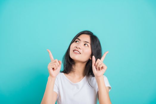Portrait Asian beautiful young woman wearing white T-shirt standing smile white teeth, She pointing finger up and looking up, shoot photo in studio on blue background, There was copy space.