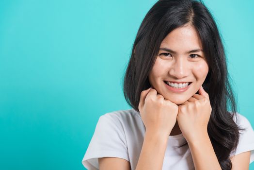 Portrait Asian beautiful happy young woman wear t-shirt glad keeps both hands under chin smiles pleasantly, on blue background, with copy space