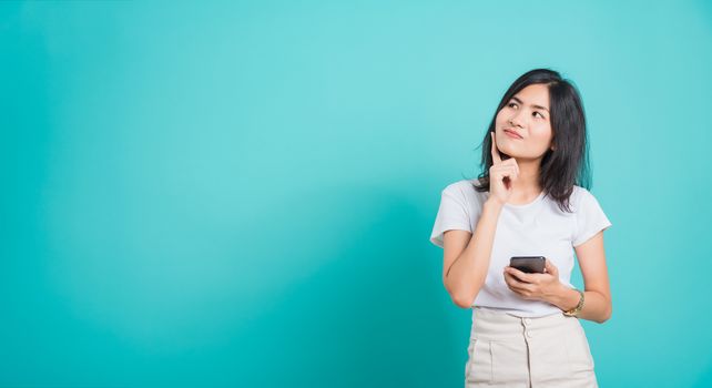 Portrait Asian beautiful happy young woman standing smile, posing using mobile phone her thinking dreaming on blue background, with copyspace