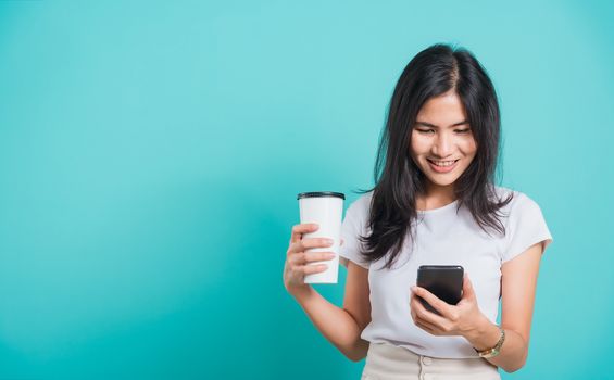 Portrait Asian beautiful happy young woman standing smile, using mobile phone her holding coffee paper cup, looking smartphone on blue background, with copyspace