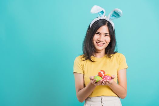 Portrait Asian beautiful happy young woman smile white teeth wear yellow t-shirt standing with bunny ears and holding Easter eggs looking to camera, on a blue background with copy space