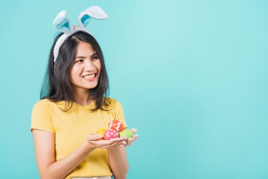 Portrait Asian beautiful happy young woman smile white teeth wear yellow t-shirt standing with bunny ears and holding Easter eggs looking to space, on a blue background with copy space