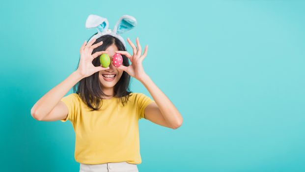 Portrait Asian beautiful happy young woman smile white teeth wear yellow t-shirt standing with bunny ears and holding Easter eggs near the eye, on blue background with copy space