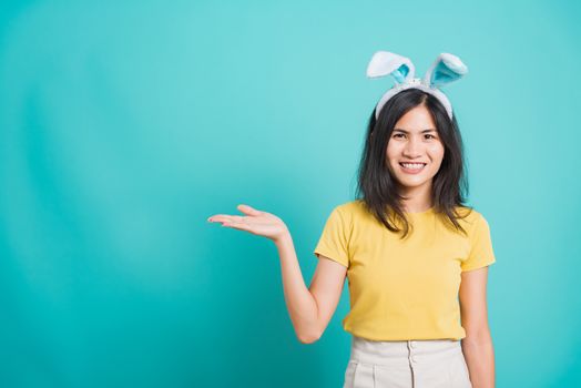 Portrait Asian beautiful happy young woman smile white teeth wear yellow t-shirt standing with bunny ears showing hand bank product her looking camera, on blue background with copy space