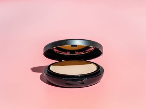 Compact powder on pink background. Female pressed powder in ajar opened black plastic case with mirror, copy space for text or design. Hard light