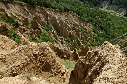 Fragment of the famous Stob’s Pyramids with unusual shape red and yellow rock formations, green bushes and trees around, west share of Rila mountain, Kyustendil region, Bulgaria, Europe