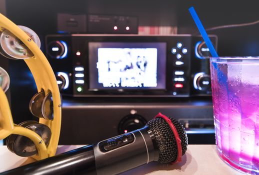 Black microphone in karaoke club, with remote controller, melon and strawberry soda drinks, yellow tambourine and screen for singing music on stage party.