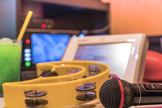 Black microphone in karaoke club, with remote controller, melon and strawberry soda drinks, yellow tambourine and screen for singing music on stage party. 