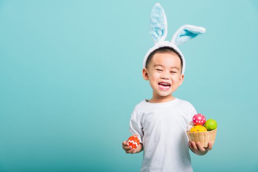 Asian cute little child boy smile beaming wearing bunny ears and a white T-shirt, standing to hold a basket with full Easter eggs. And other hand holds an easter egg on blue background with copy space