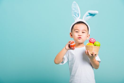 Asian cute little child boy smile beaming wearing bunny ears and a white T-shirt, standing to hold a basket with full Easter eggs. And other hand holds an easter egg on blue background with copy space