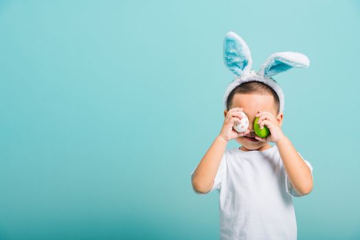 Asian cute little child boy smile beaming wearing bunny ears and a white T-shirt, standing to holds colored easter eggs instead of eyes on blue background with copy space for text