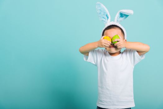 Asian cute little child boy smile beaming wearing bunny ears and a white T-shirt, standing to holds colored easter eggs instead of eyes on blue background with copy space for text