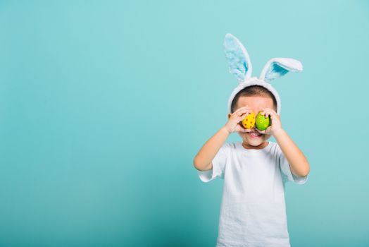 Asian cute little child boy smile beaming wearing bunny ears and a white T-shirt, standing to holds colored easter eggs instead of eyes on blue background with copy space for text