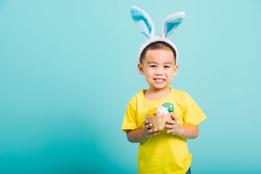 Asian cute little child boy smile beaming wearing bunny ears and a yellow T-shirt, standing to hold a basket with full Easter eggs on blue background with copy space