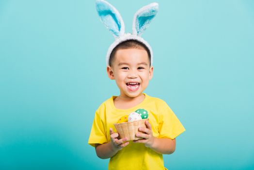 Asian cute little child boy smile beaming wearing bunny ears and a yellow T-shirt, standing to hold a basket with full Easter eggs on blue background with copy space